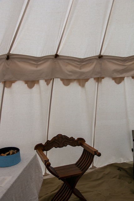 The inside of a white tent. An olive-colored tarp is laid on the floor with an ornate wooden stool in the lower frame. A table with a white sheet is the left of the stool with a blue tin on top.