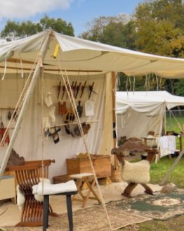 A large white sutler wedge tent set up among other similar tents in a grassy area. The tent has a few carpets laid out on the floor with stools set up on top of it. There are poles hung up on the wall of the tent to show several various items on display.