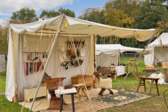 A large white sutler wedge tent set up among other similar tents in a grassy area. The tent has a few carpets laid out on the floor with stools set up on top of it. There are poles hung up on the wall of the tent to show several various items on display.