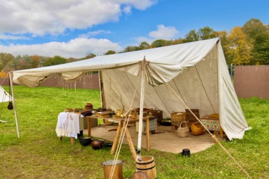 A large white sutler wedge tent set up in an open grassy space with a brown fence behind it. The tent is serving as an outdoor kitchen with a table set up and several pots and containers on top of and underneath. Behind it are several baskets and storage boxes. In front of the tent is a barrel and another pot.