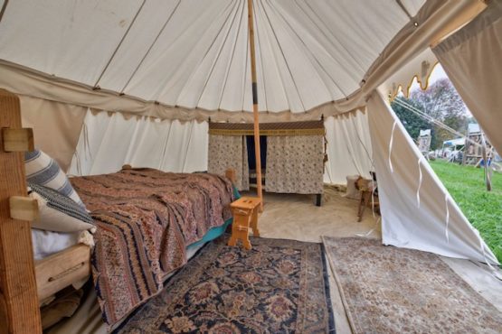The inside of a large white round end tent. A pole is pushing the roof up high to give the tent a larger interior. A tarp is laid on the floor with two carpets on top of it. A bed is set up on the left side of the tent with a stool beside it. On the opposite side is the flap entrance of the tent left open.