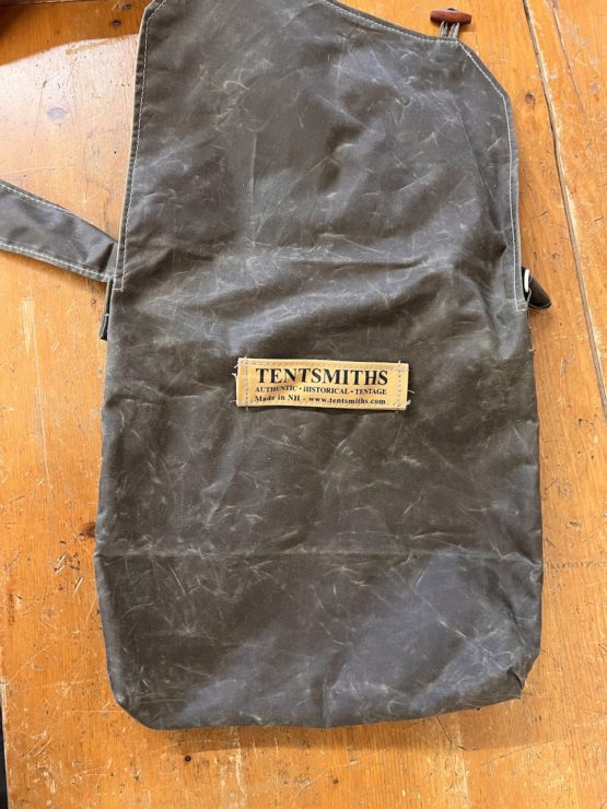 The back of a satchel bag laid flat on a wooden surface that has a tag stiched into it. It reads 'Tentsmiths. Authentic Historical Tentage. Made in NH. www.tentsmiths.com"