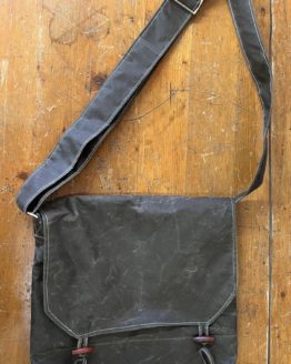 A black satchel bag closed and laid flat on a wooden surface with the shoulder strap stretched above it.