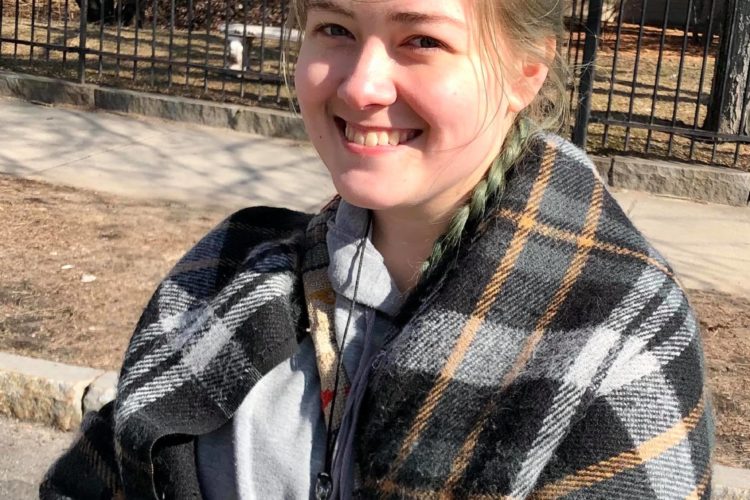 A woman smiling while standing in a street. She's wearing a blue shirt and draped in a plaid blanket. Her hair is dyed green and braided into a ponytail that hangs over her shoulder. Behind her is a metal fence separating the walkway from someone's backyard.