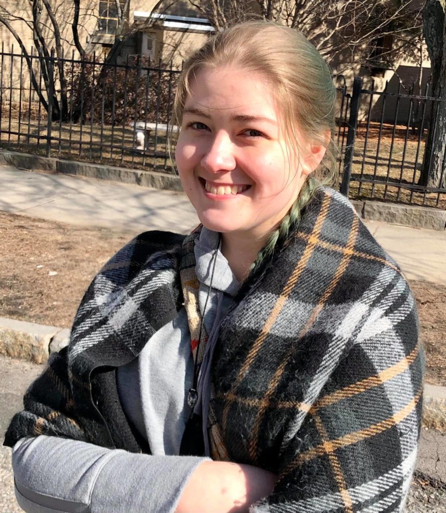 A woman smiling while standing in a street. She's wearing a blue shirt and draped in a plaid blanket. Her hair is dyed green and braided into a ponytail that hangs over her shoulder. Behind her is a metal fence separating the walkway from someone's backyard.