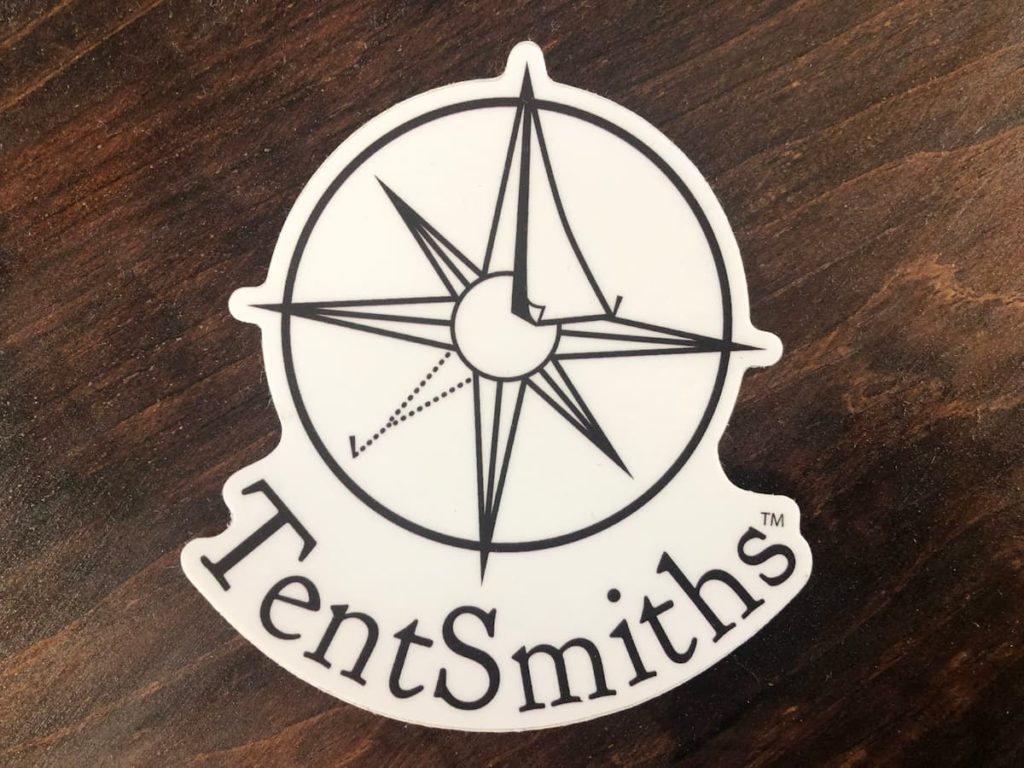 Our Very First Swag! A white sticker on a wooden surface. In black ink, it reads TentSmiths and curves around underneath the company logo.