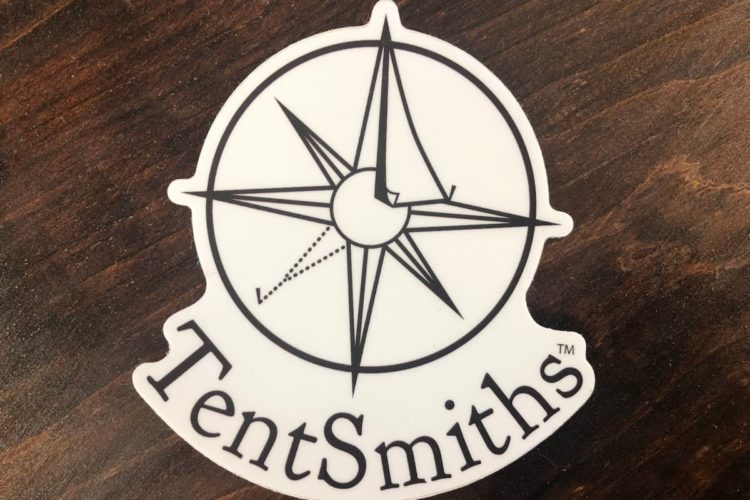 A white sticker on a wooden surface. In black ink, it reads TentSmiths and curves around underneath the company logo.