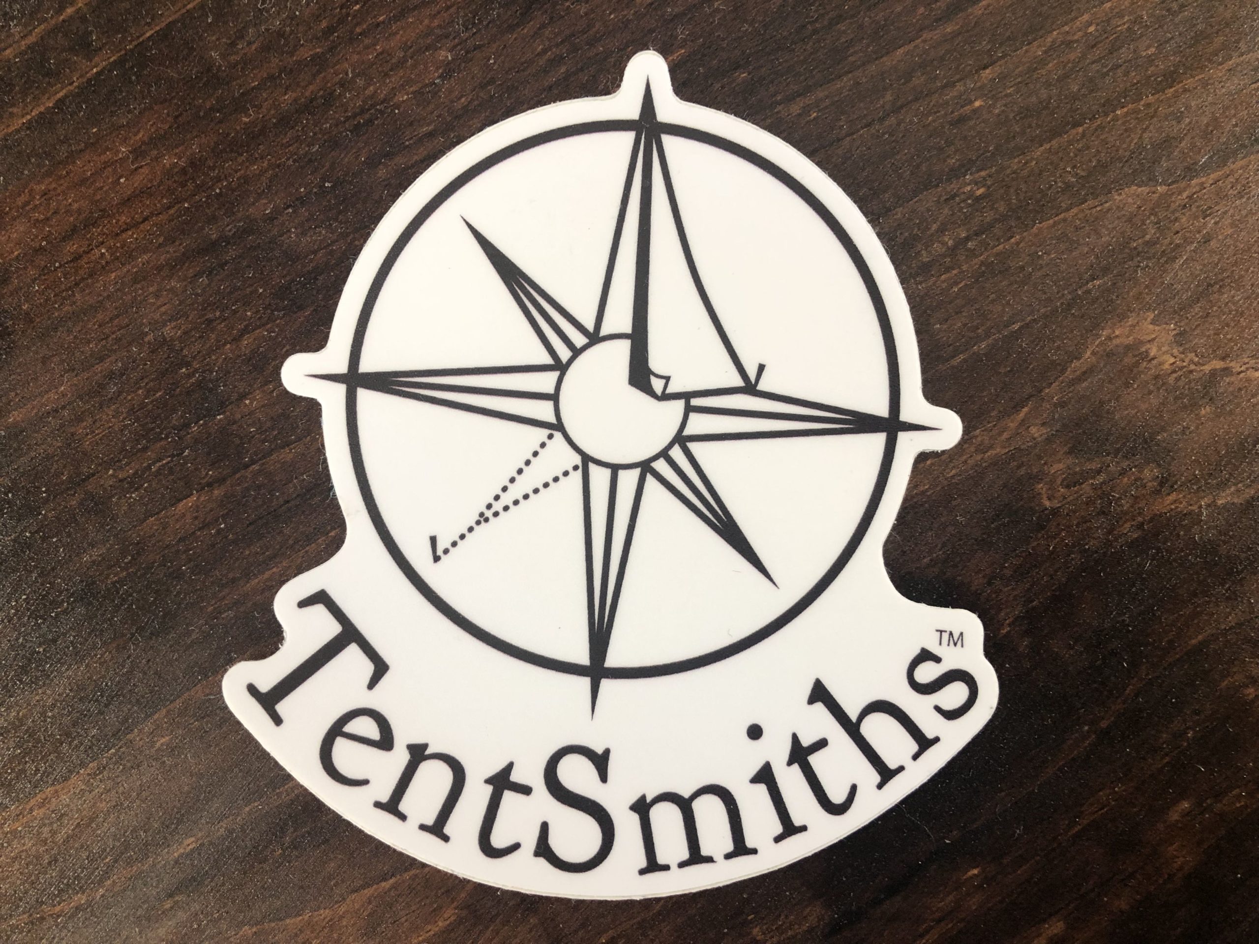 Our Very First Swag! A white sticker on a wooden surface. In black ink, it reads TentSmiths and curves around underneath the company logo.