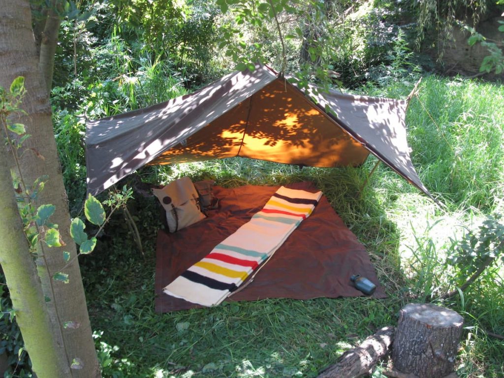 An oilskin tarp is hung from a tree to provide a cover over a comfortable campsite. A brown blanket is laid flat on the ground with another blanket, white with multi-color stripes on both ends, folded on top of it. The campsite is located in a grassy spot with trees and bushes providing a backdrop.