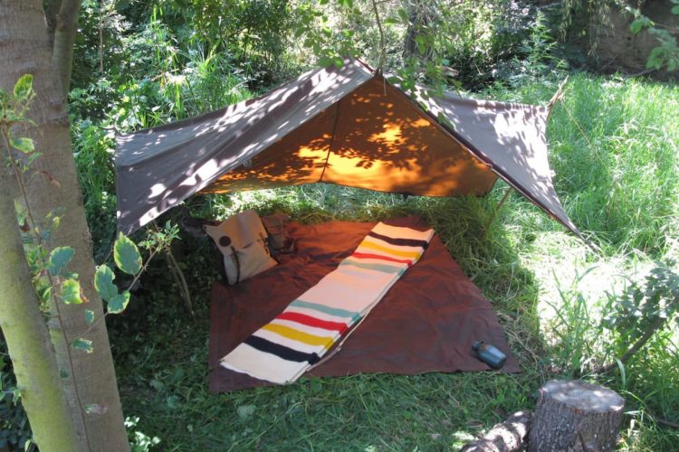 An oilskin tarp is hung from a tree to provide a cover over a comfortable campsite. A brown blanket is laid flat on the ground with another blanket, white with multi-color stripes on both ends, folded on top of it. The campsite is located in a grassy spot with trees and bushes providing a backdrop.