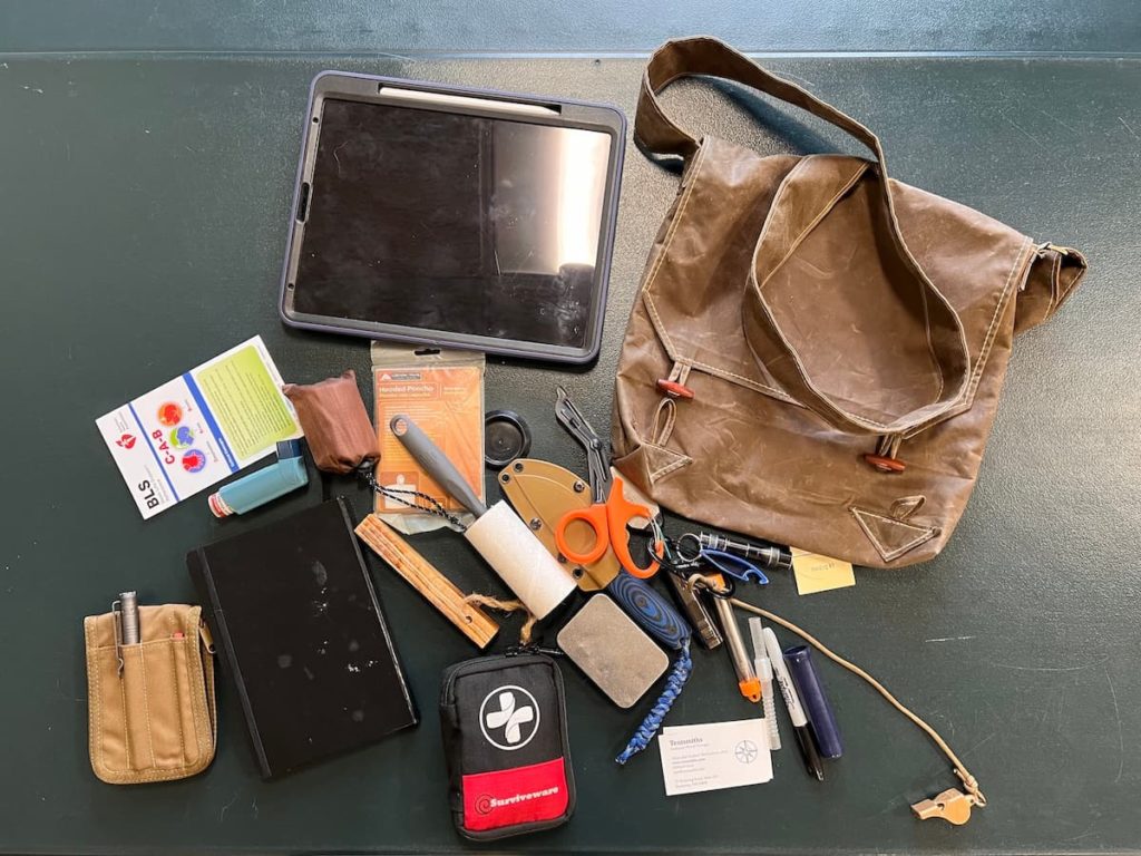The contents of a bag laid out on a green surface; a personal tablet with a pen, a pamphlet, an astham inhaler, a small pouch, a packaged hooded poncho, a lint roller, a book, a pen and pencil container made of fabric, a first aid kit, a couple of business cards, a sharpie, a pair of sccissors, a flashlight, a bottle opener, a whistle, and other items for every day outdoor journeying. The bag is in the top right of the pile, and is meant to imply that all of the items will fit inside.
