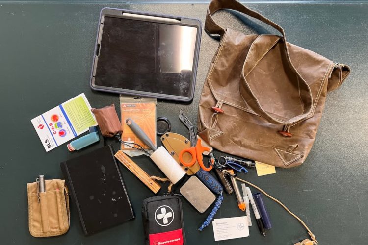 The contents of a bag laid out on a green surface; a personal tablet with a pen, a pamphlet, an astham inhaler, a small pouch, a packaged hooded poncho, a lint roller, a book, a pen and pencil container made of fabric, a first aid kit, a couple of business cards, a sharpie, a pair of sccissors, a flashlight, a bottle opener, a whistle, and other items for every day outdoor journeying. The bag is in the top right of the pile, and is meant to imply that all of the items will fit inside.